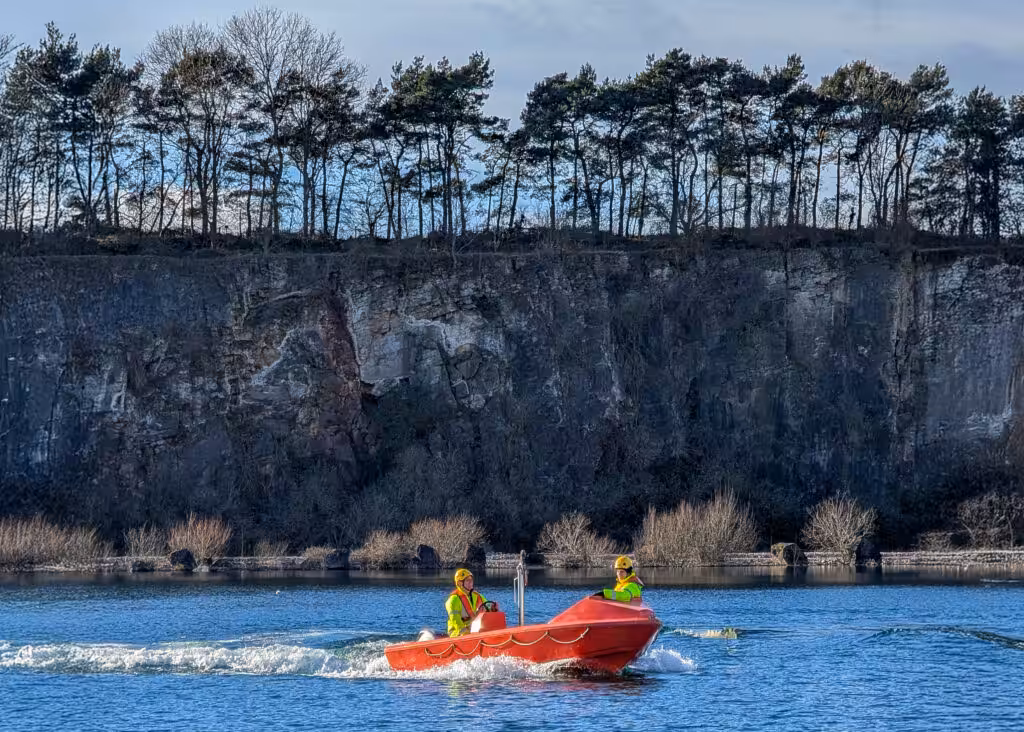 A rescue boat training exercise by South West Maritime Academy taking place in the water body at Cromhall Quarry. The cliff face in the background a mix of grey and tan colours, showing distinct layers, deep vertical cracks, and a rough, weathered texture.