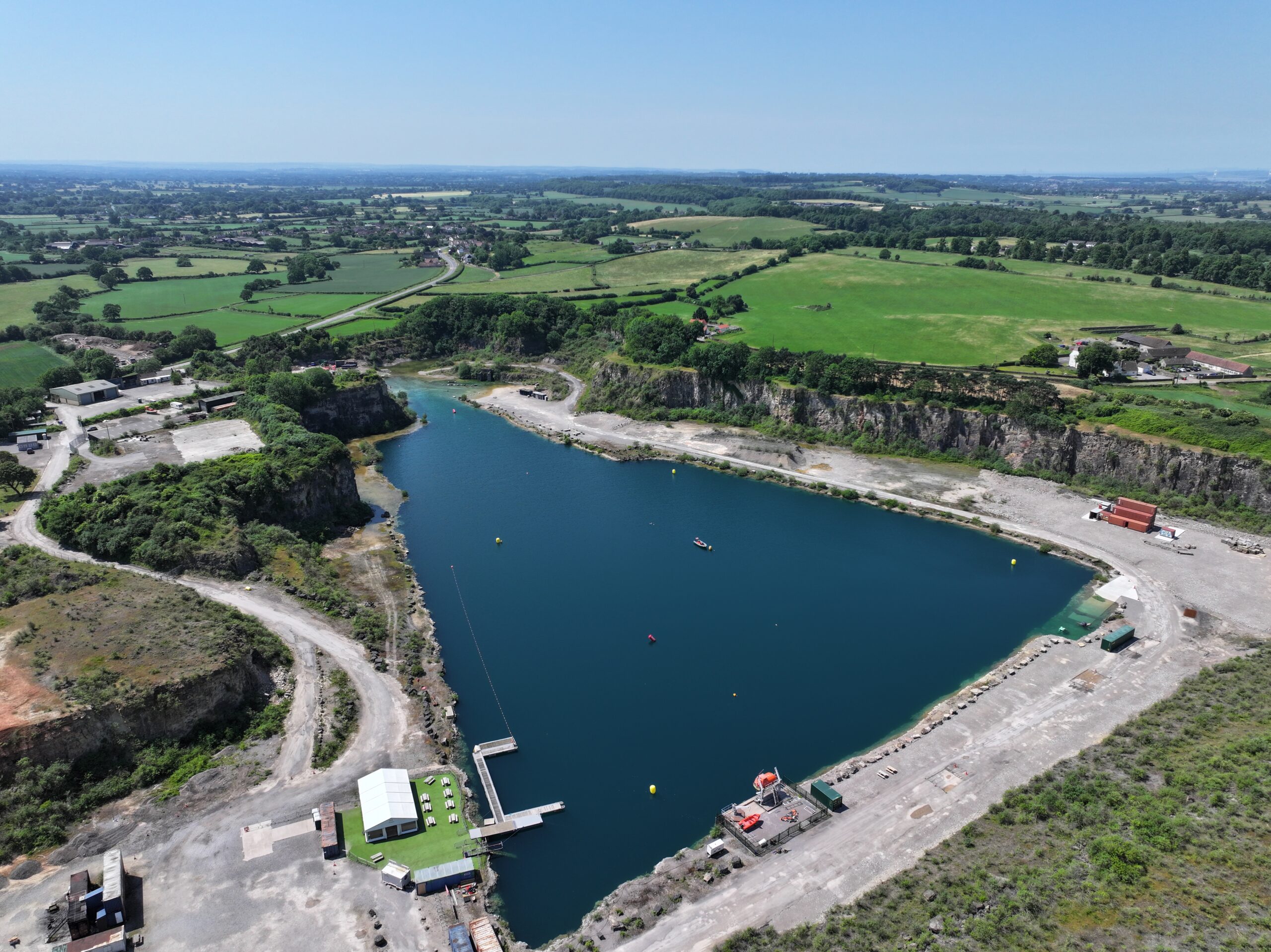 A wide, aerial photograph of Cromhall Quarry on a bright day. Sheer, layered rock cliffs surround a large, calm body of deep blue water. Green vegetation grows on the upper edges of the cliffs and fields surrounding the quarry.